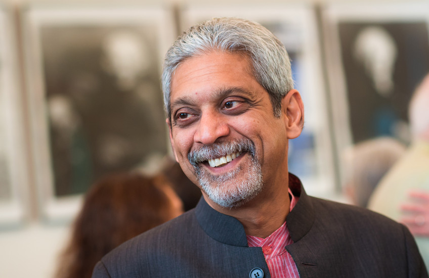 A silver-haired man, wearing a black jacket and a red shirt, smiles at someone off camera.