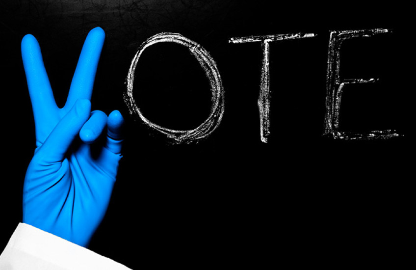 Photo of chalkboard with the word vote - the V is being made by the hand of a person with surgical gloves on.