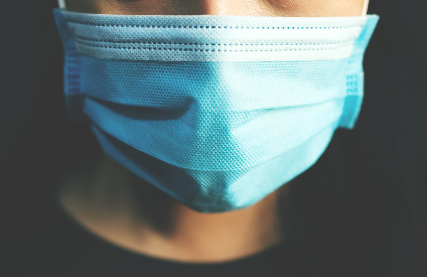 Closeup of the face of a person wearing a paper medical mask in front of a dark background.