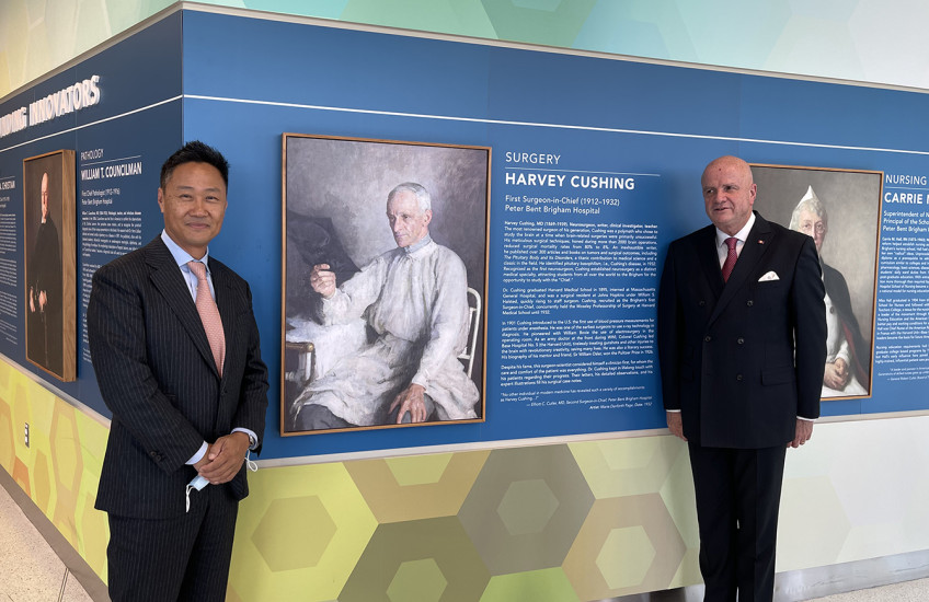 In a hallway painted with colorful geometric patterns, two men in suits stand on either side of a portrait of a man in surgical gowns reviewing an X-ray of a patient’s skull. The portrait is labeled “Harvey Cushing.”
