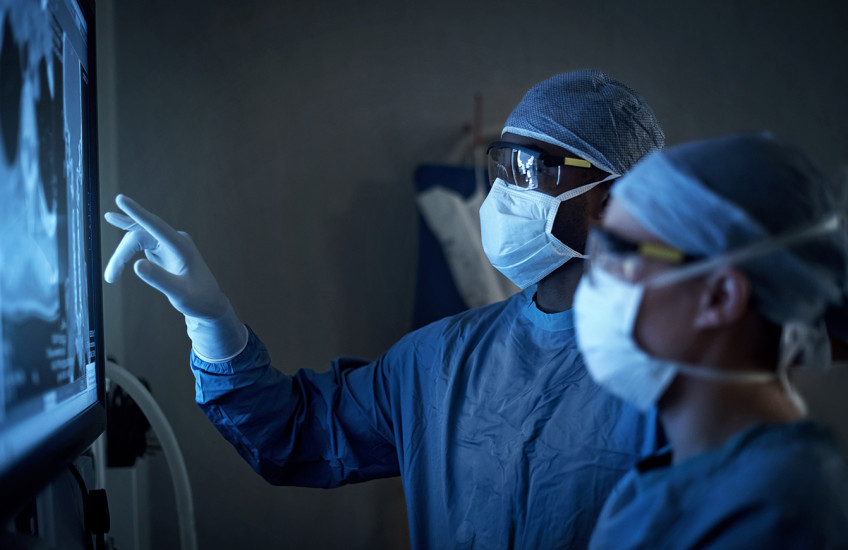 Two figures wearing surgical masks, gowns, and caps examine a backlit X-ray image