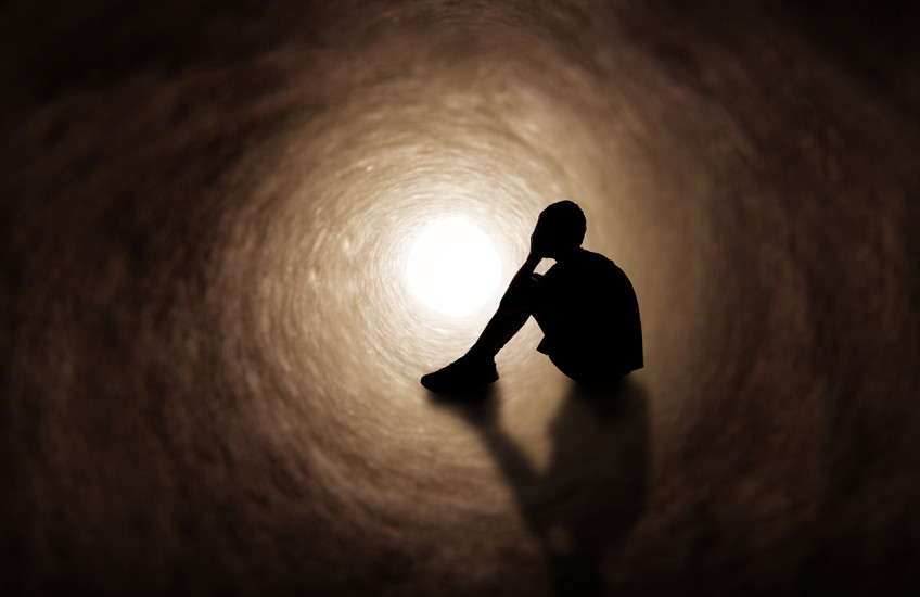 The silhouette of a young person in shorts and t-shirt covering their face as they sit in a dark tunnel.