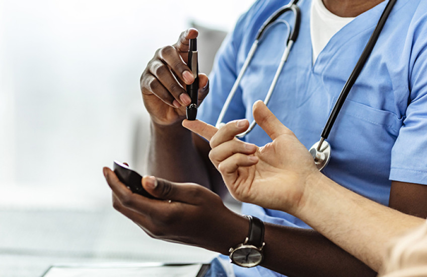 Health care provider in blue scrubs with stethoscope and darker skin tests the blood sugar of a light-skinned patient 