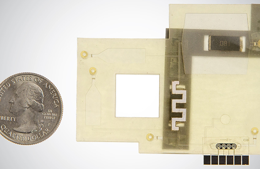 Close-up view of a flat, plastic-looking device with many square angles, an opening in the center, and a microfluidic strip. It is about 4 times the size of a quarter coin shown to the side.