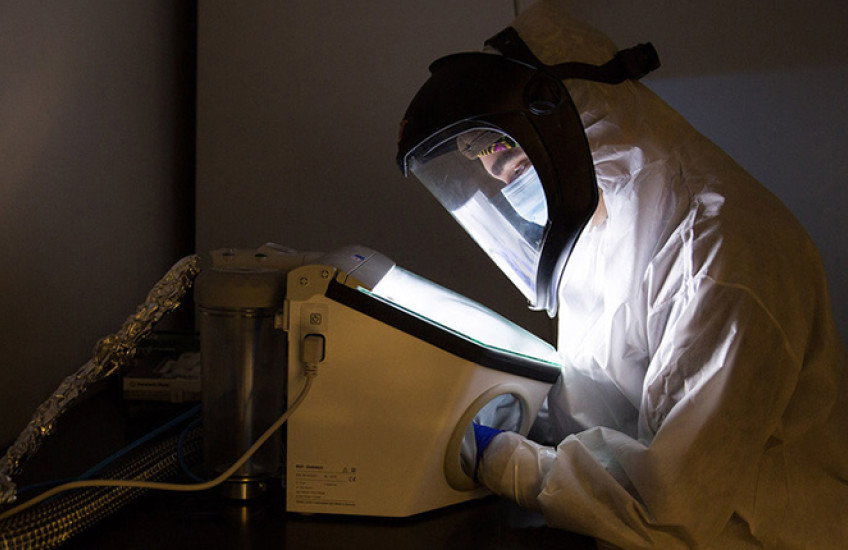 Person in a full-body clean suit and masked hood leans over a lit display in a dark room