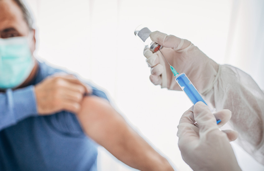 Gloved hands prepare a needle for an injection while the patient rolls up their sleeve.