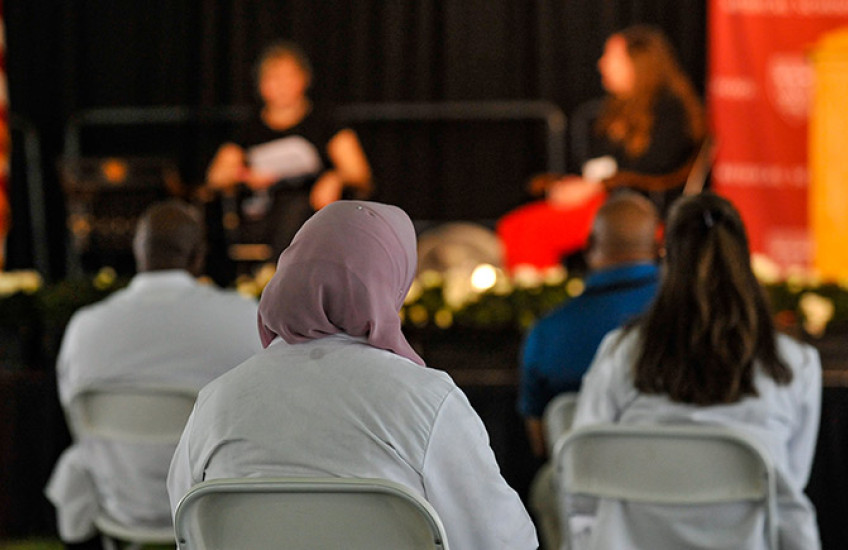 Three people in white coats in focus in audience watch a female patient and female doctor on stage out of focus
