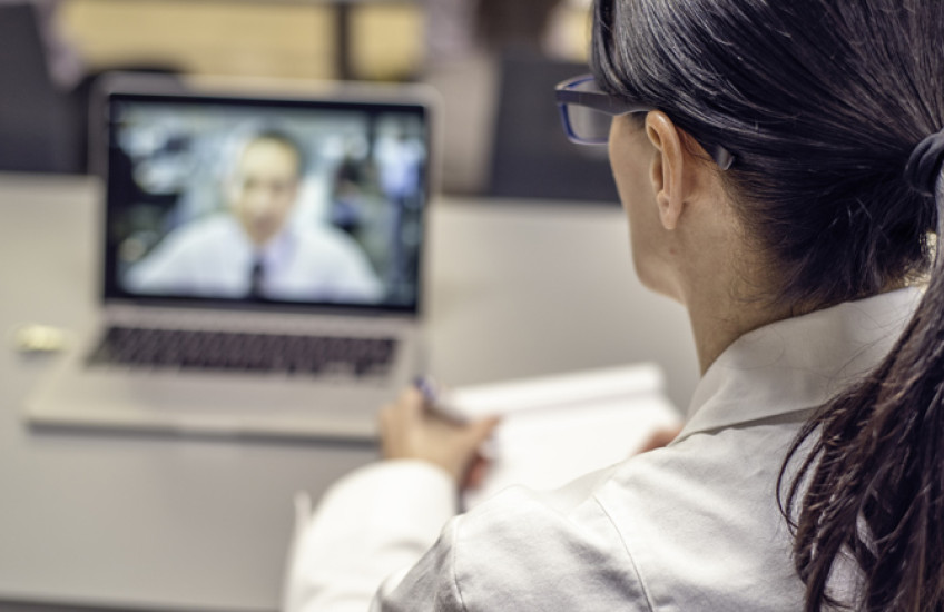Looking over a doctor's shoulder, we see a patient video conferencing on the doctor's laptop.