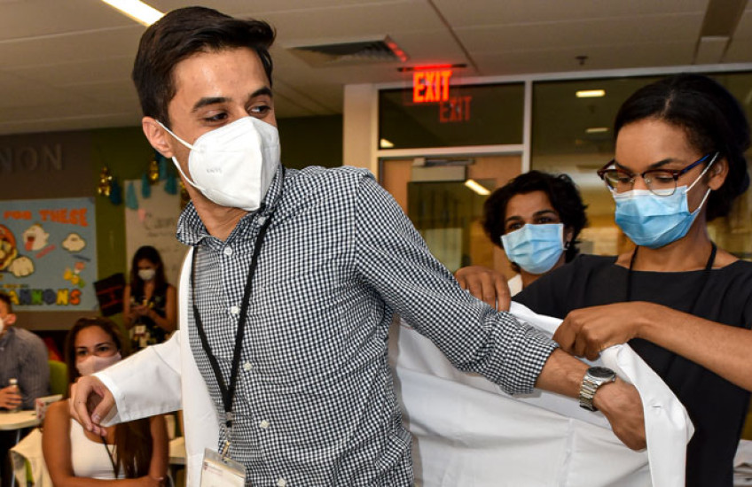 Male student in check shirt wearing a mask puts on a white coat with help of woman