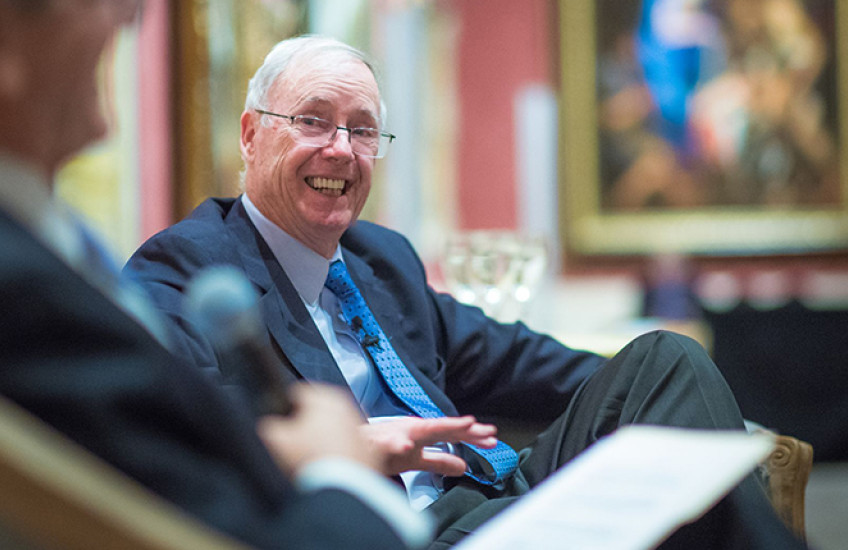 Candid photo of a gray-haired man in glasses grinning in a chair while being interviewed