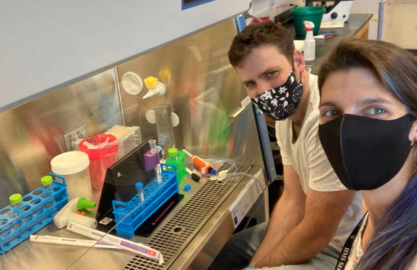 Two people in masks smile beside a hooded lab enclosure