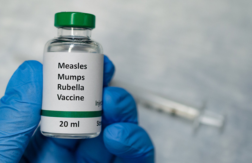 Gloved hand holds a vial labeled "Measles mumps rubella vaccine 20 milliliters" with syringe in background