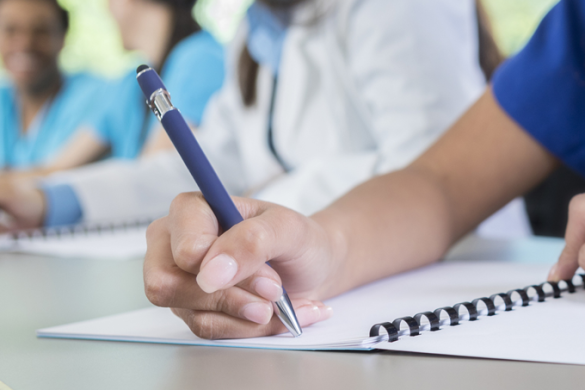 A close-up of a forearm with a pen in the hand, writing in a notebook, with other people blurred in the background.