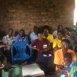 A group of adults seated closely together inside a simple brick-walled room listen attentively as two women stand at the front speaking. The audience includes men and women of different ages, some holding notebooks or bags, creating the feel of a community meeting or training session.