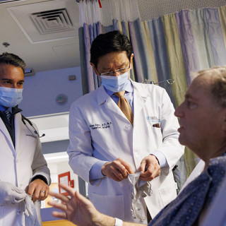A patient in a hospital bed talks to two surgeons in doctors’ coats and masks.