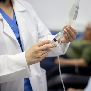 A health care professional administers a medication in a syringe connected to a hanging IV bag beside a seated patient.