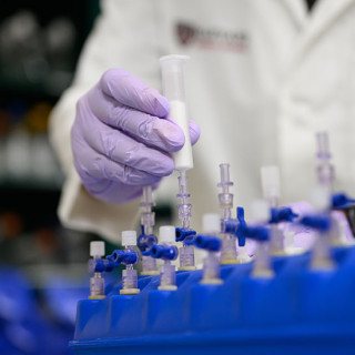A researcher wearing purple gloves handles a syringe connected to laboratory equipment with multiple valves and tubes arranged in a blue rack