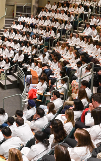 Wide angle view of 200 students in an auditorium wearing white coats with six people sitting on a panel at the front of the room.