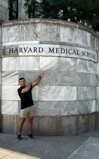 A first-year student grins and points at the HMS sign on the marble entrance to the Quad