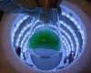 A flask of green liquid held by a gloved hand is silhouetted at the top of a cylinder with rings of lights