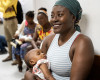 A seated woman in a sleeveless shirt and a headscarf holds a sleeping baby in the foreground, while more people sit in a line behind them in a waiting room. 