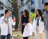 Four medical students, two in white coats, smiling and walking together on a sunny day with a marble columned building in background.