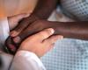 Image of a doctor's hands holding a black hospital patient's hands.  No faces.