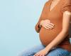 Cropped stock image of a woman with light brown skin sitting in a hair with a hand on her pregnant belly. A bandage on her arm indicates a recent COVID vaccination.