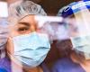 The faces of two people in medical scrubs, protective masks, and head gear seen through a reflection-covered window.