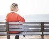 Woman with light hair and orange sweater sits alone on a bench overlooking water