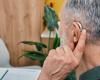 back of the head of a white grey-haired man holding a finger to his ear that has a hearing aid
