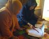 Two women, one in orange and one in blue seated on a colorful rug look at documents together.