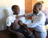 A teenage boy sits on an examination table in a clinic as a man listens to his heart with a stethoscope.