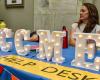 A women sits behind a table with lit up signage letters the spell out OCCME