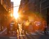 A group of people cross a street at a crosswalk, backlit by the glow of the sun and surrounded by a halo of rainbowed lens flare.
