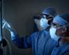 Two figures wearing surgical masks, gowns, and caps examine a backlit X-ray image