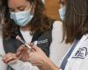 Two women in white coats and surgical masks hold syringes labeled "COVID-19 vaccine"