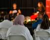 Three people in white coats in focus in audience watch a female patient and female doctor on stage out of focus
