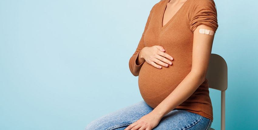 Cropped stock image of a woman with light brown skin sitting in a hair with a hand on her pregnant belly. A bandage on her arm indicates a recent COVID vaccination.