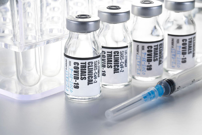 vials labeled "clinical trials COVID 19" in front of test tube rack, with syringe in foreground