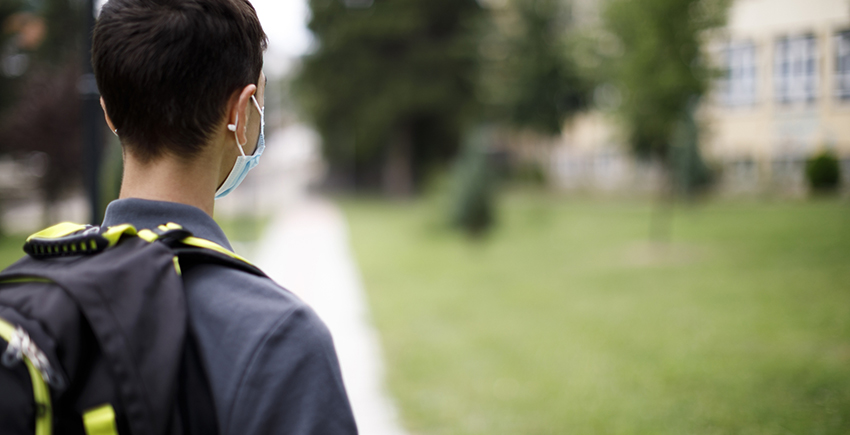 Rear view of a child with backpack and mask on facing a schoolyard