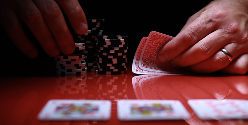 an unseen person at a shiny gambling table holds unknown cards while picking up a few poker chips from a stack. on the table lie three blurry cards face-up.