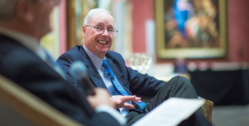 Candid photo of a gray-haired man in glasses grinning in a chair while being interviewed