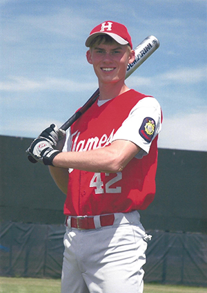 A smiling young man poses in a baseball uniform, holding a bat over one shoulder