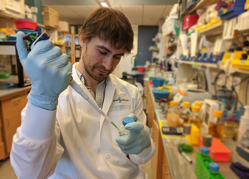 A young man wearing a lab coat pipettes green liquid into a small container while standing in a lab