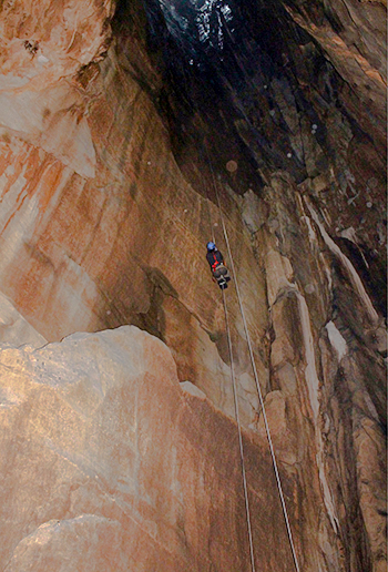 Woman in blue helmet climbs up a rope, dwarfed by tall rock face, with light at top in the distance
