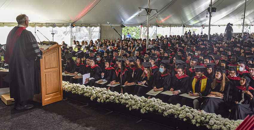 View of student and guest crowd under a tent with over-the-shoulder shot of Dean Daley at podium