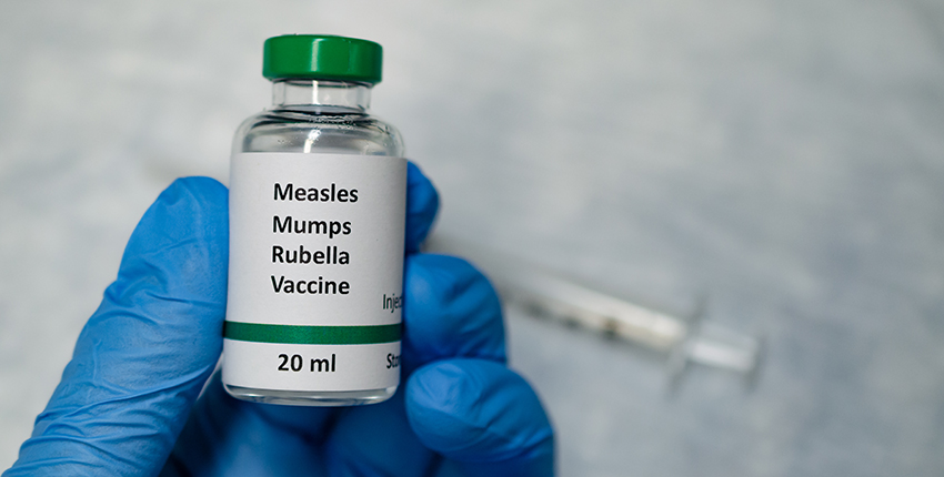 Gloved hand holds a vial labeled "Measles mumps rubella vaccine 20 milliliters" with syringe in background
