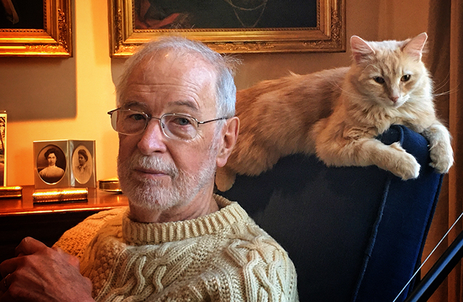 professor in sweater with cat on back of armchair, in softly lit living room with portraits on wall resembling a museum