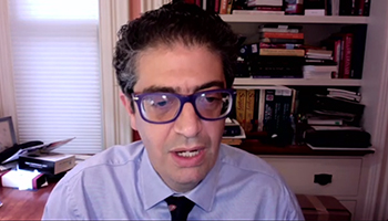 Screen grab of man in glasses speaking in front of a bookshelf, looking slightly off to the side